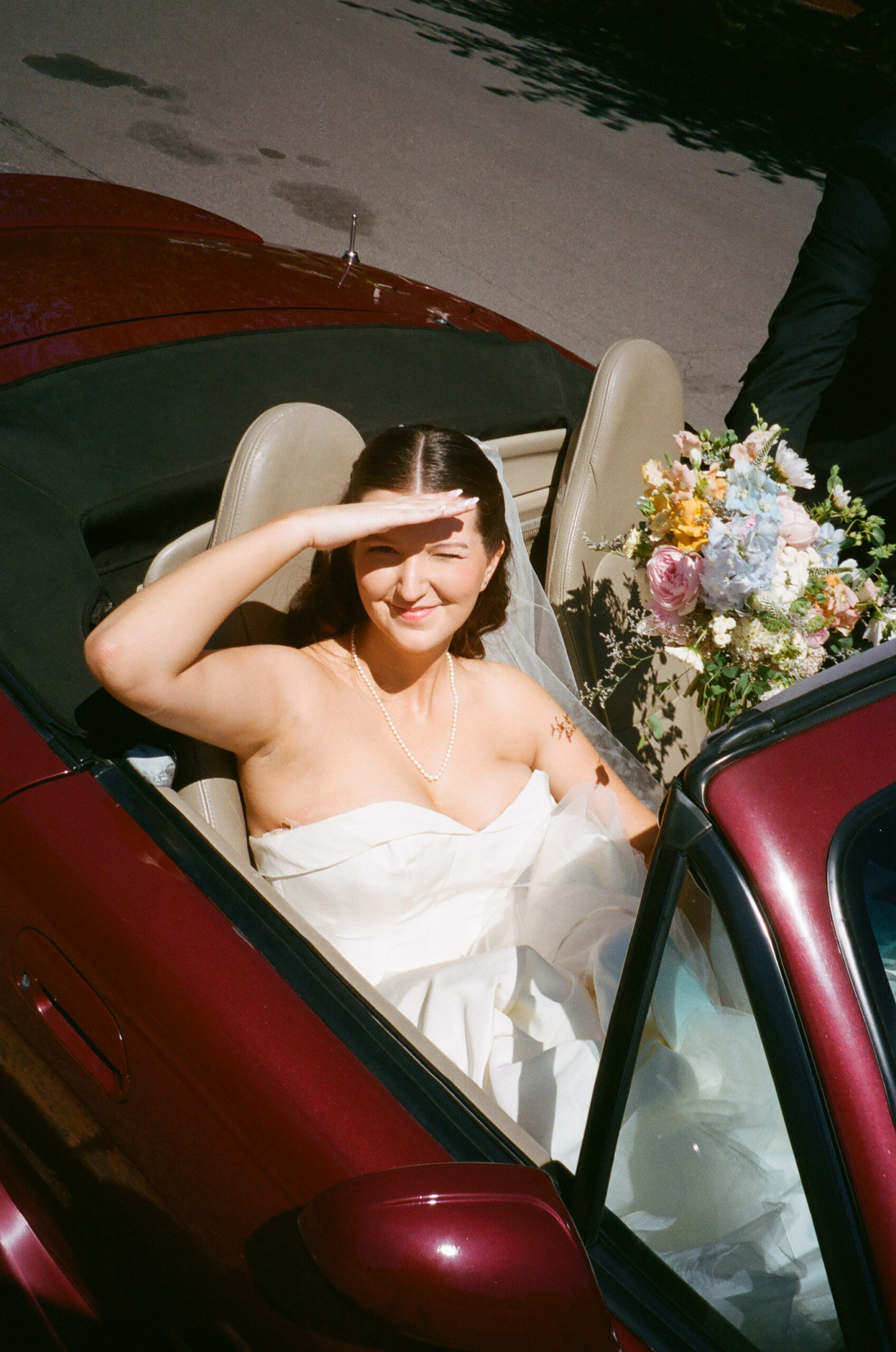 Film photo of bride covering her eyes away from the sun sitting in her getaway car