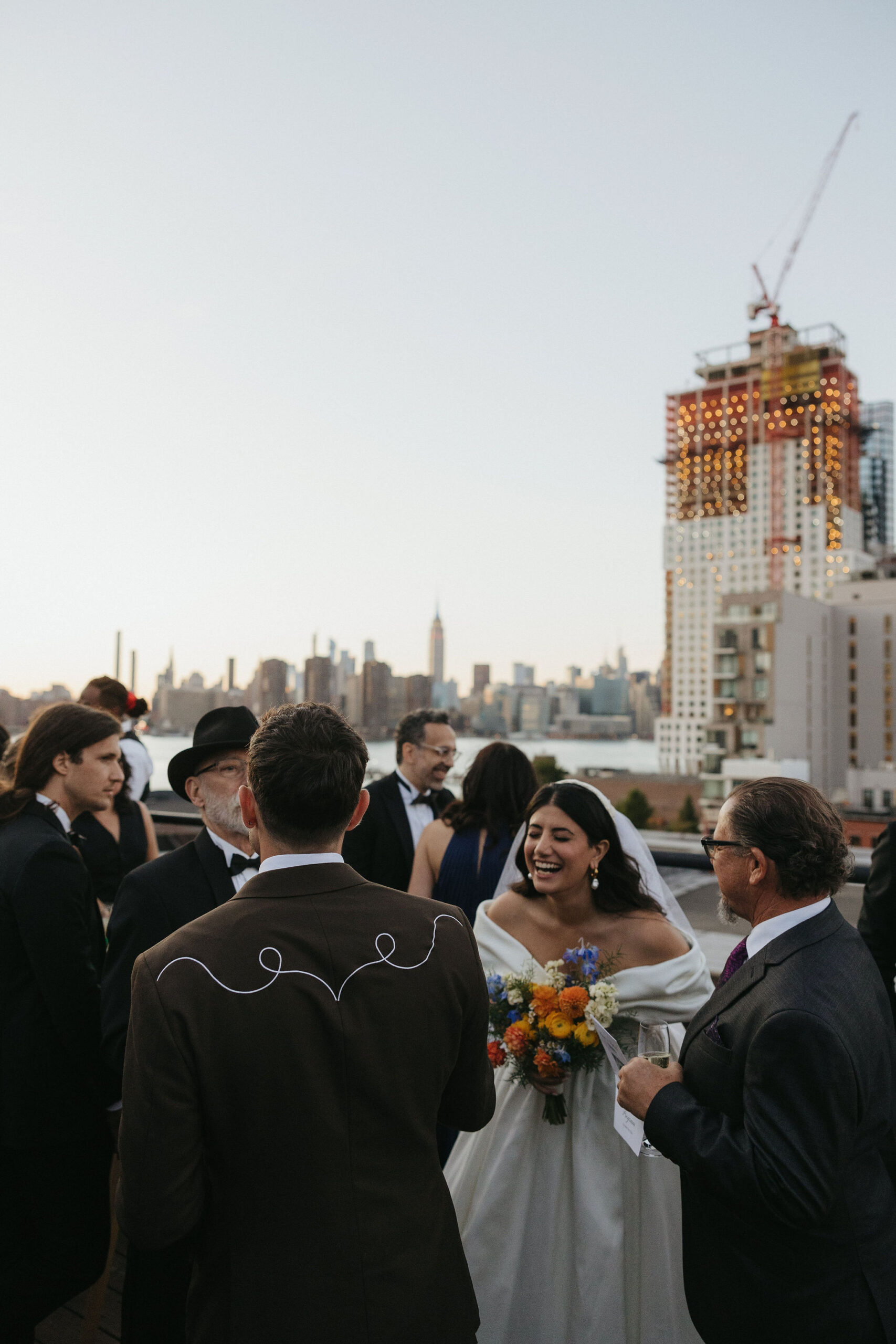 Greenpoint Loft Wedding reception rooftop gathering with guests laughing, the bride holding a bright bouquet of flowers.
