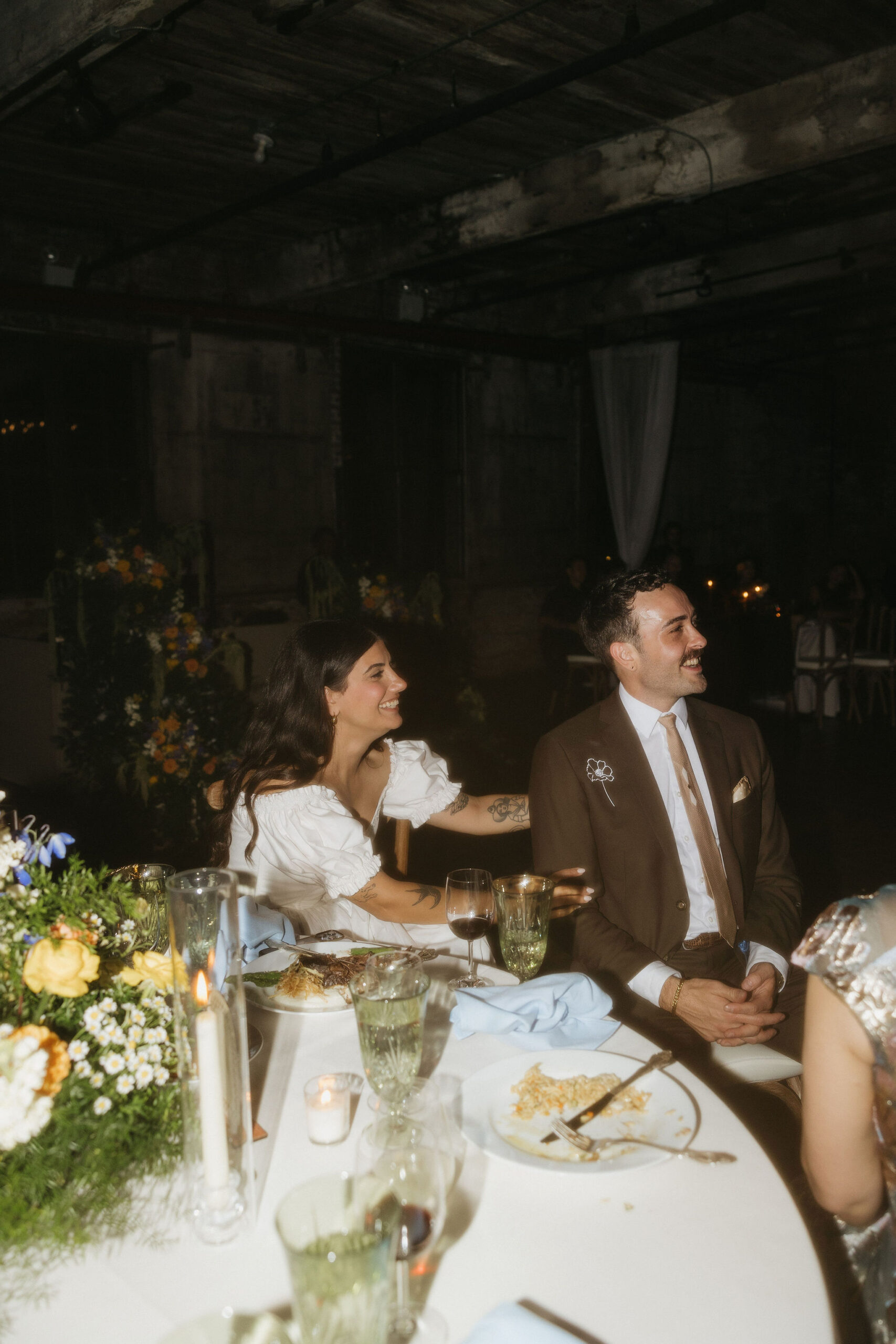 bride and groom sitting together at a beautifully decorated reception table during a Greenpoint Loft Wedding, with the bride in a white dress and the groom smiling in a brown suit.