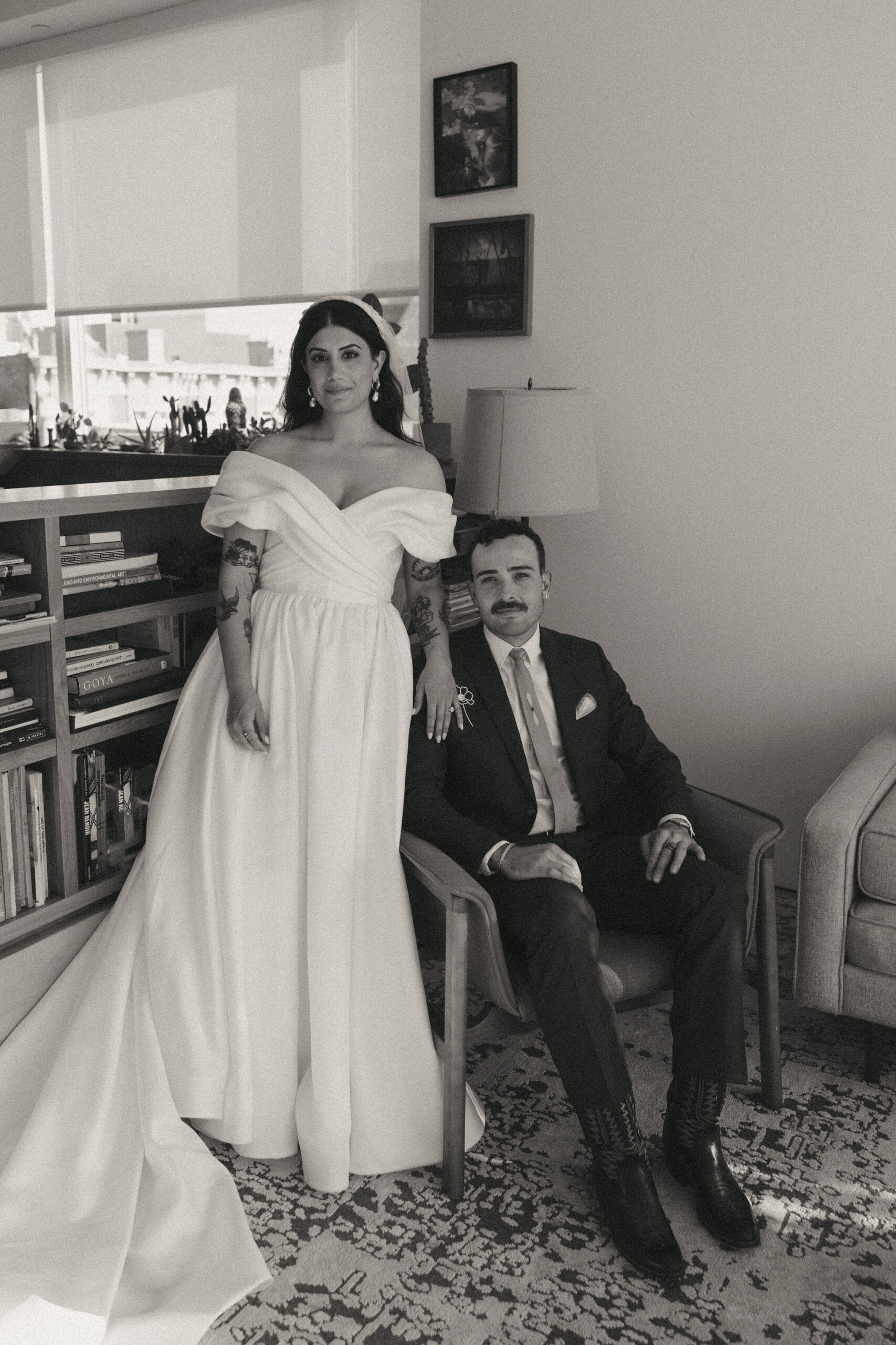 A black and white film wedding photo of a couple posing indoors, featuring the bride in an elegant off-shoulder gown.